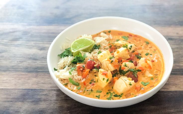 a white bowl filled with soup and rice on top of a wooden table next to a slice of lime