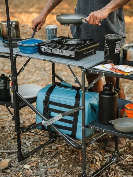 a man cooking food on top of a metal table in the middle of a forest