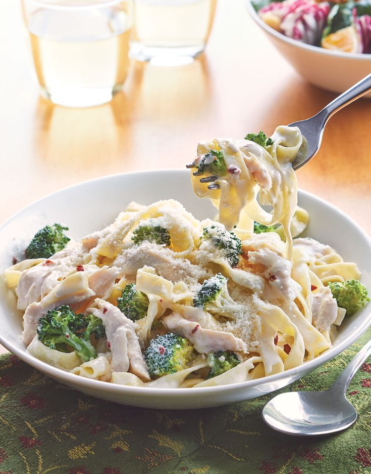 a bowl of pasta with chicken and broccoli on the table next to two glasses of water