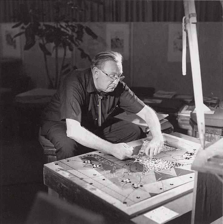 an old man sitting at a table working on a piece of art with beads and scissors