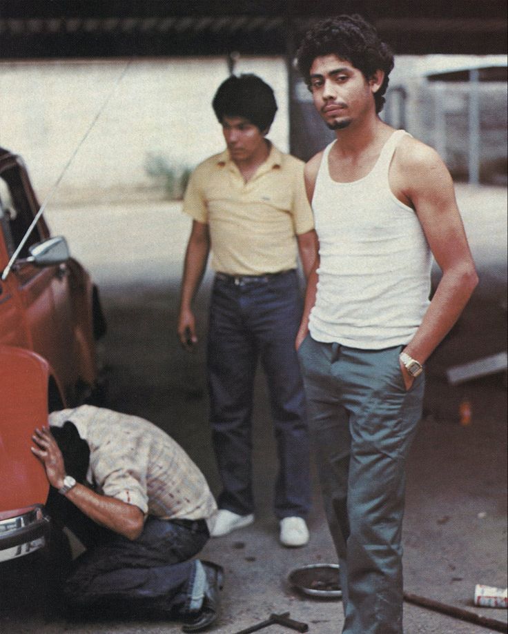 two men standing next to an old red car with the hood open and one man fixing it