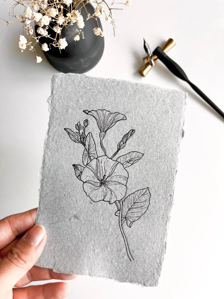 a person holding up a piece of paper with a drawing on it and flowers in the background