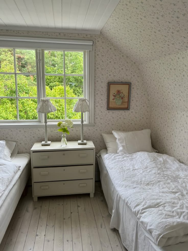 two twin beds in a small room with white walls and wood flooring, along with a window