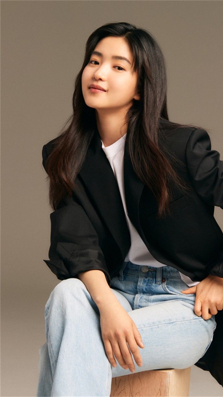 a woman sitting on top of a wooden stool wearing a black blazer and jeans