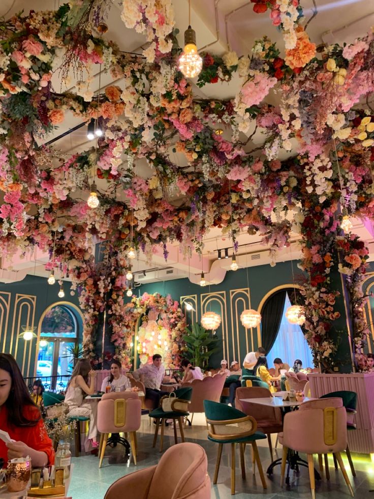 people are sitting at tables in a restaurant with flowers hanging from the ceiling above them