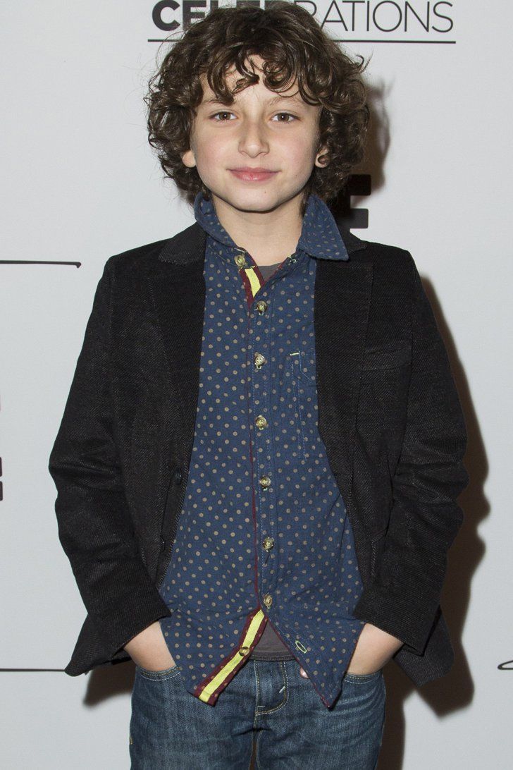 a young boy wearing a blue shirt and black blazer standing in front of a white wall