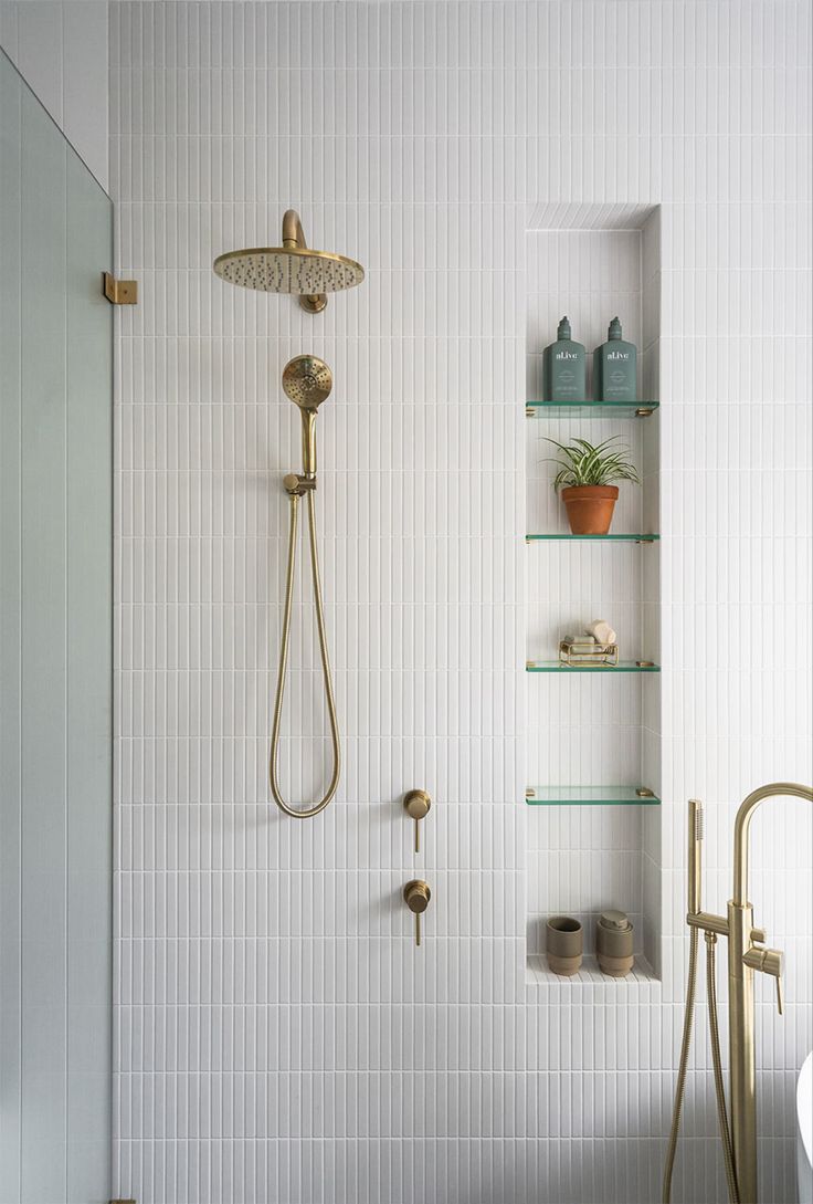 a bathroom with white tile and gold fixtures, including a shower head and shelves on the wall