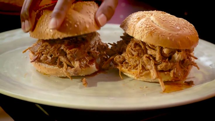 two pulled pork sandwiches on a plate being held by someone's hand over another sandwich