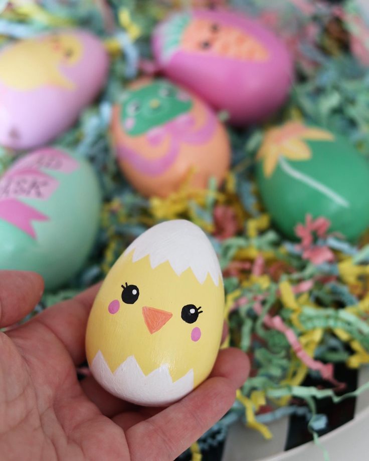 a hand is holding an egg decorated like a chick and other colorful eggs in the background