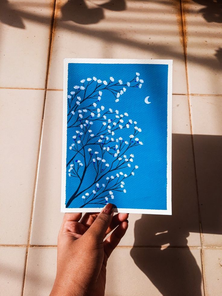 a hand holding up a blue card with white flowers on it in front of a tiled floor