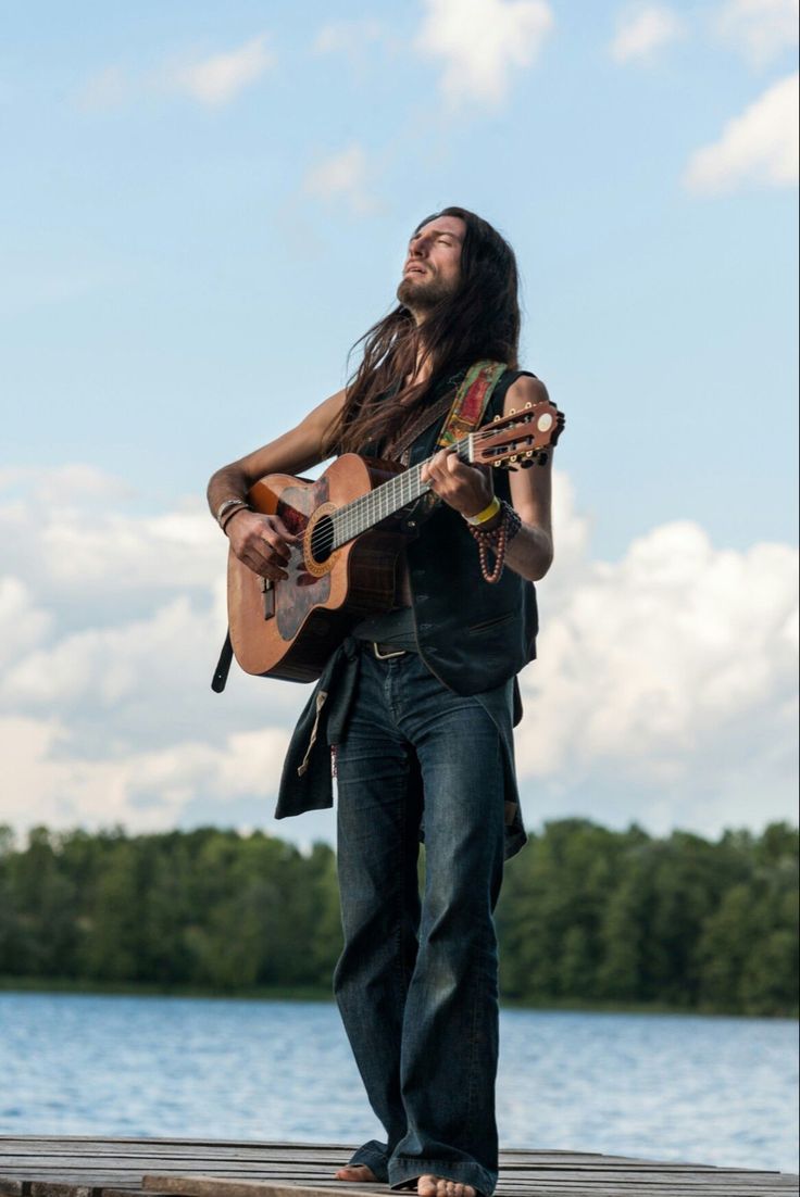 a man with long hair is playing an acoustic guitar on a dock by the water