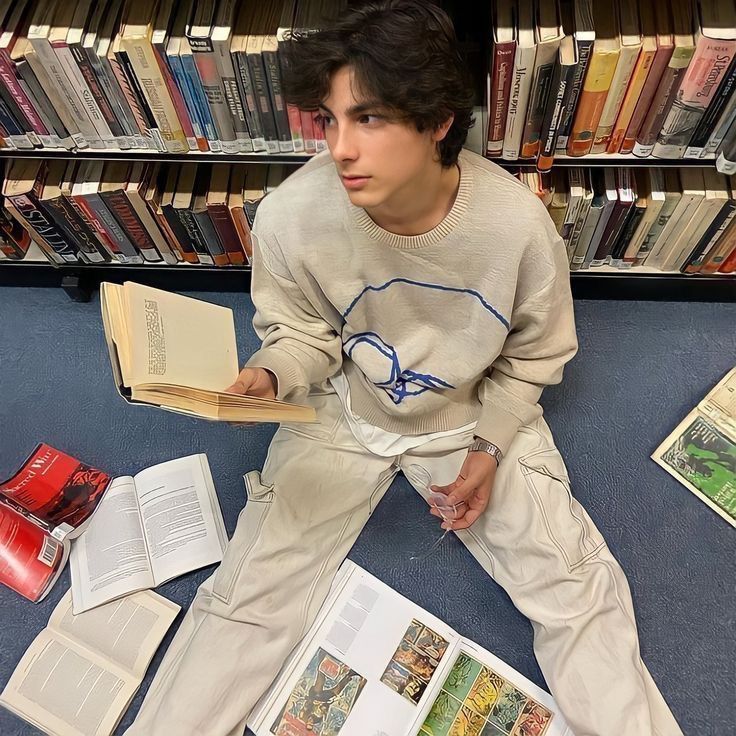 a young man sitting on the floor in front of a bookshelf holding an open book