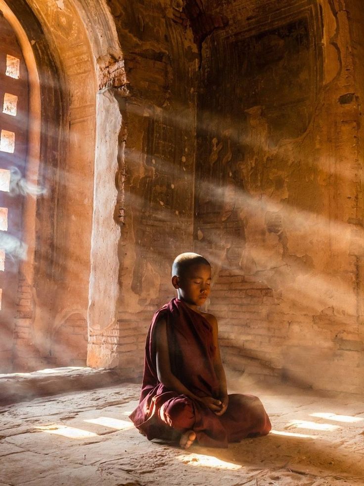 a young monk sitting on the ground in front of a stone wall with sunlight streaming through it