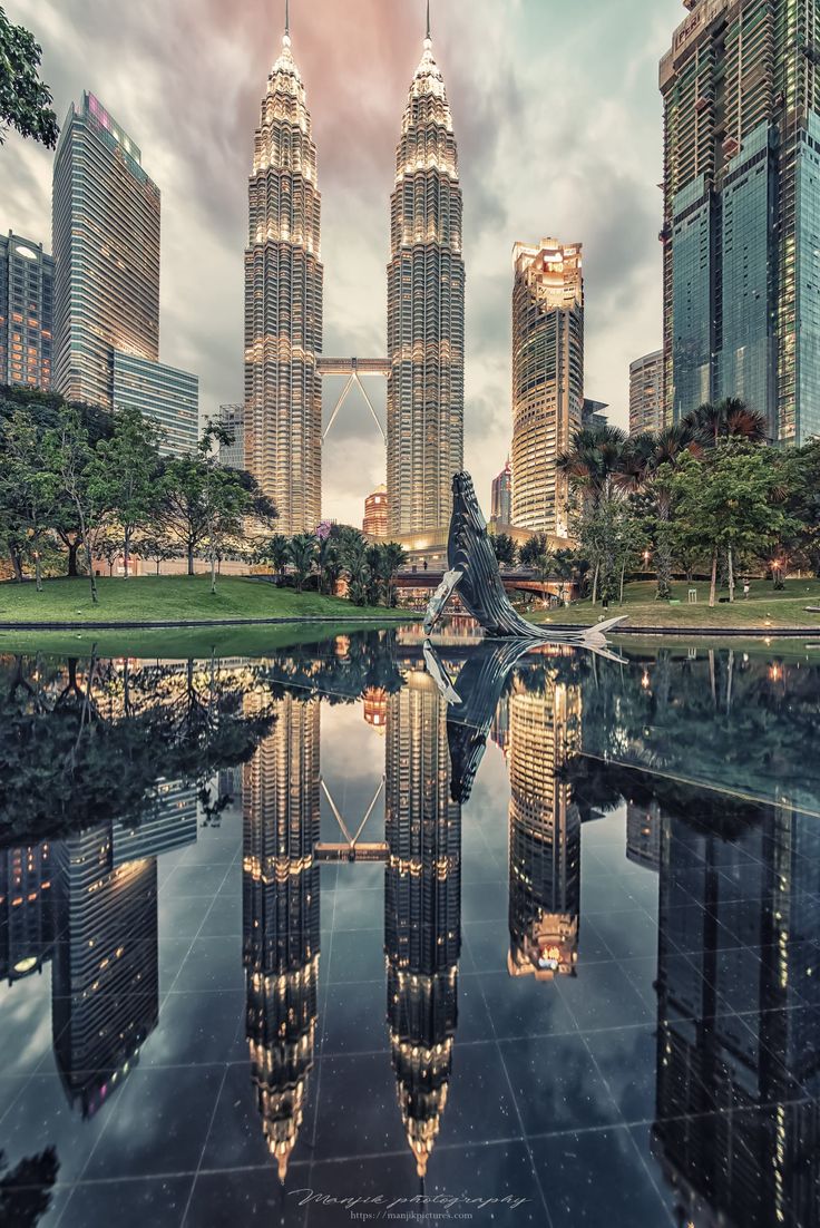 the reflection of two skyscrapers is shown in the water at dusk, with other tall buildings in the background