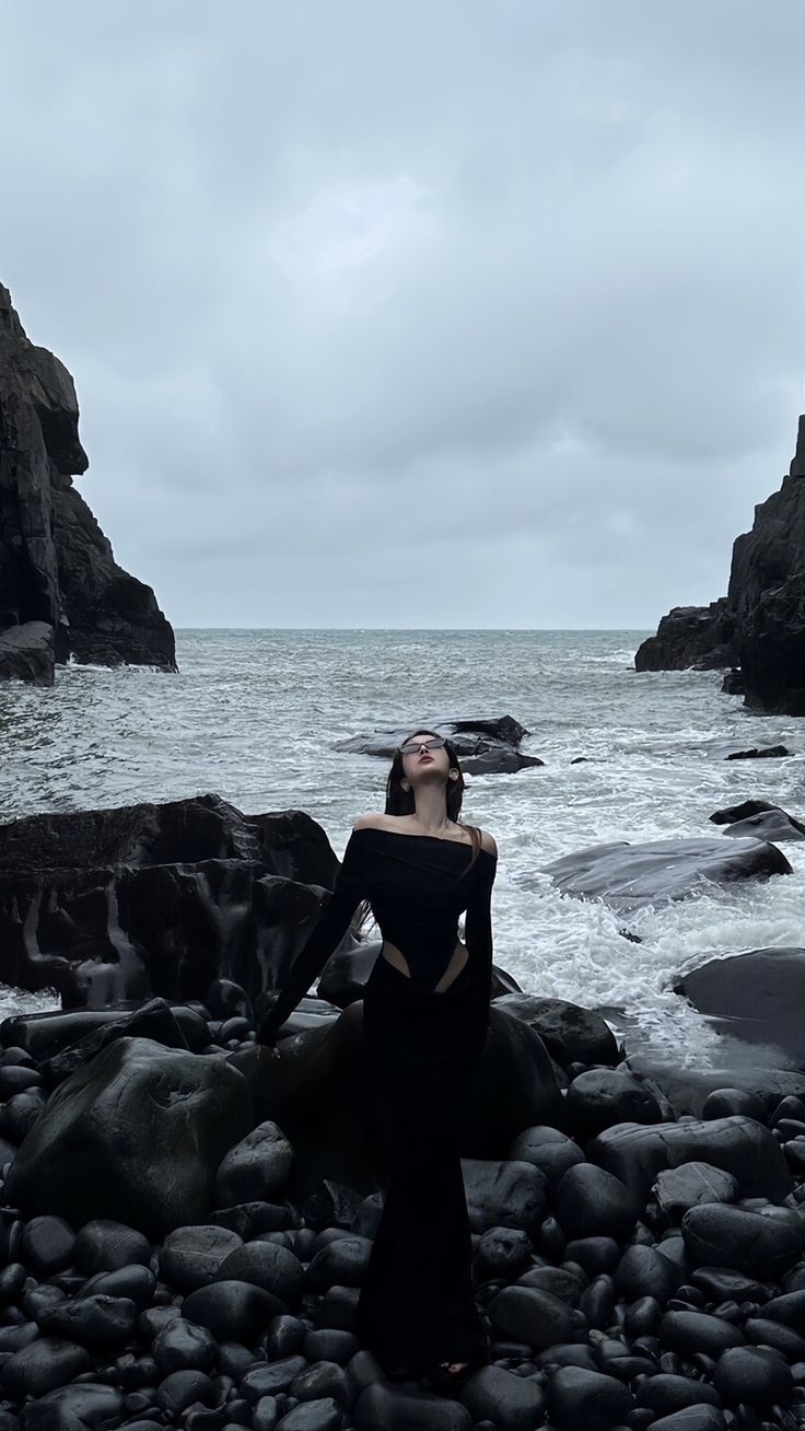 a woman standing on rocks near the ocean with her eyes closed and hands behind her head