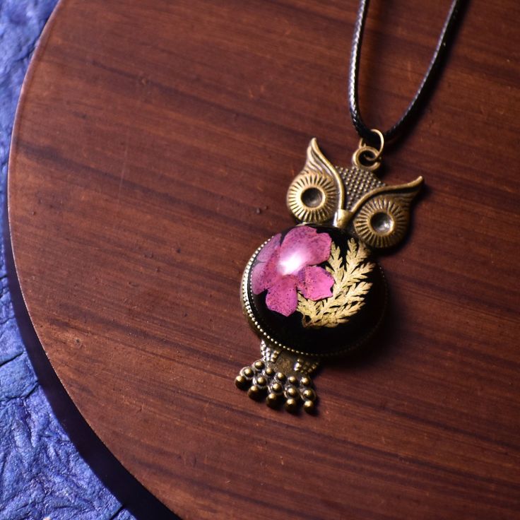 an owl necklace with a pink stone on it's face sitting on a wooden table