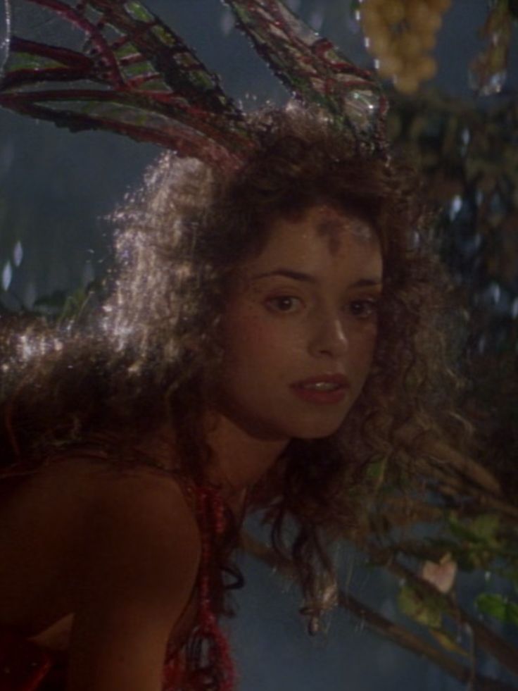 a woman with long curly hair standing in front of a tree and looking at the camera