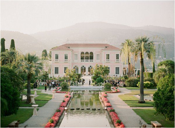 a large pink house surrounded by palm trees and water features an ornamental fountain in the foreground