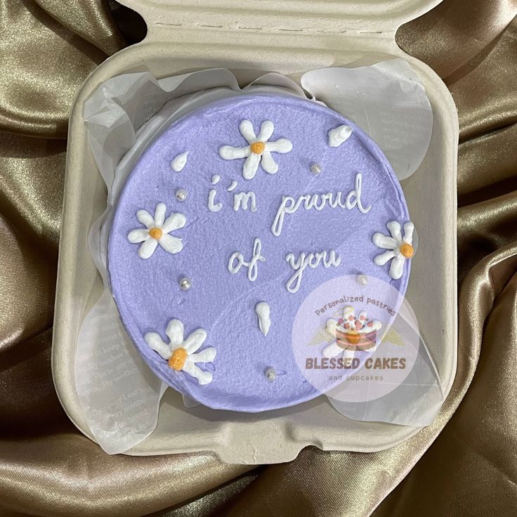 a purple cake with daisies on it in a plastic container that says i'm proud of you