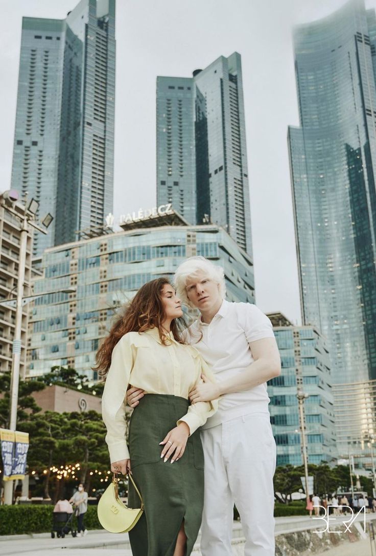 an older man and woman standing in front of tall buildings with their arms around each other