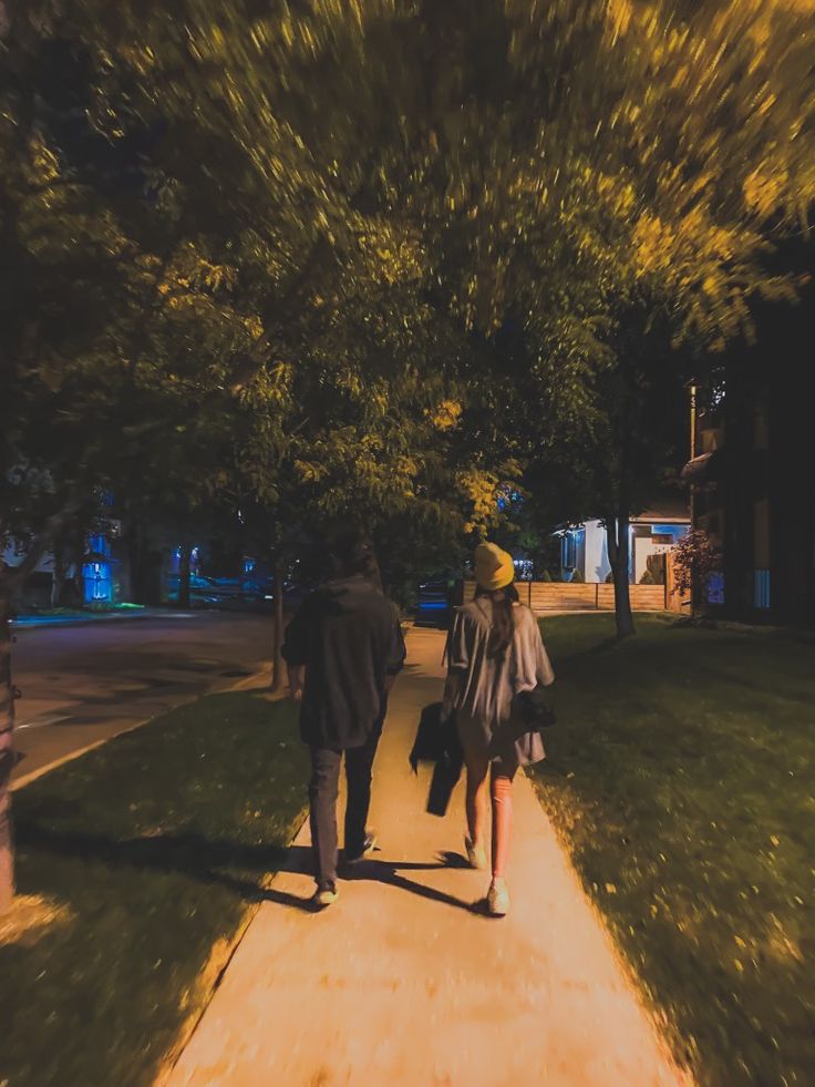 two people walking down a sidewalk in the evening with trees on either side of them