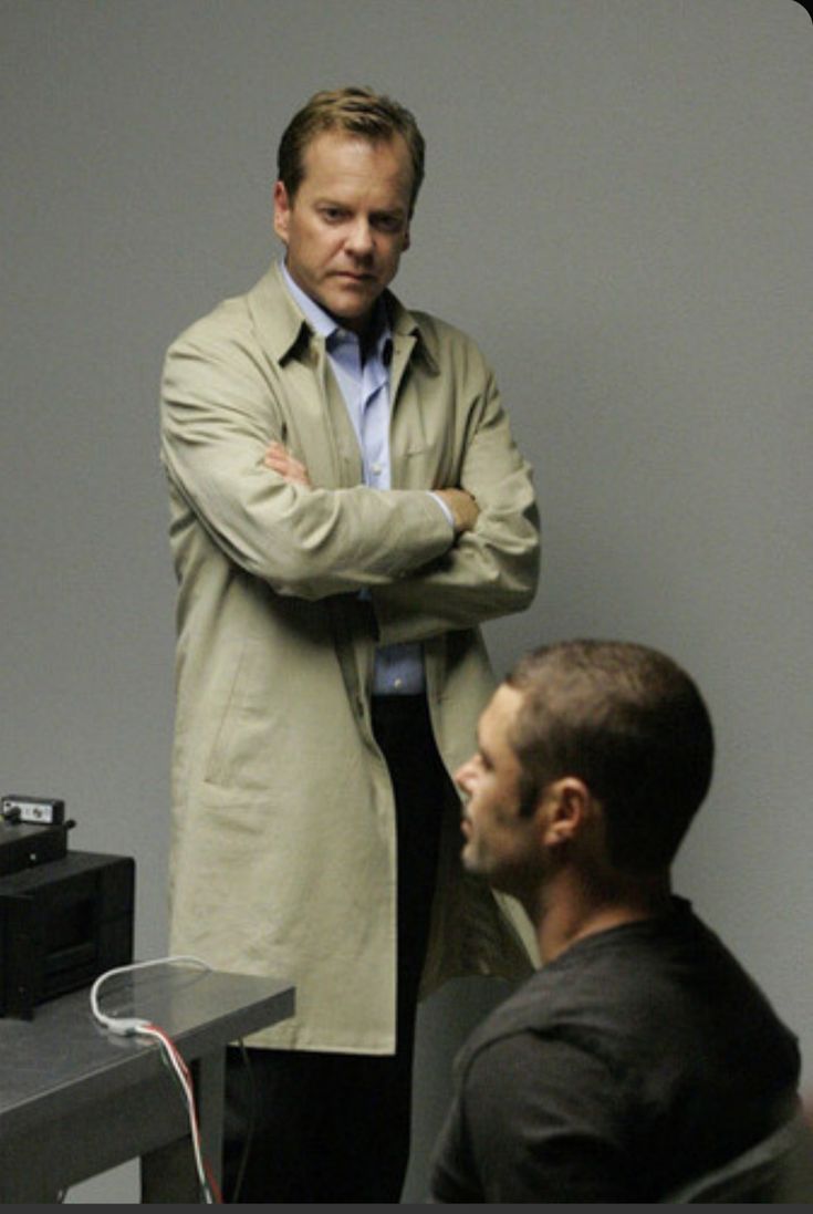 a man standing in front of a computer monitor with his arms crossed while another man looks on