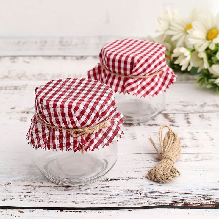 two jars with red and white gingham cloth on them sitting next to some daisies