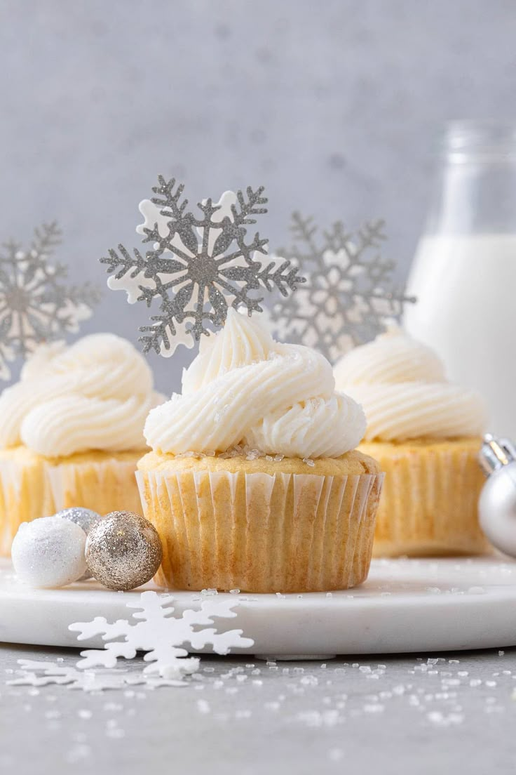 three cupcakes on a plate with snowflakes in the background and silver ornaments