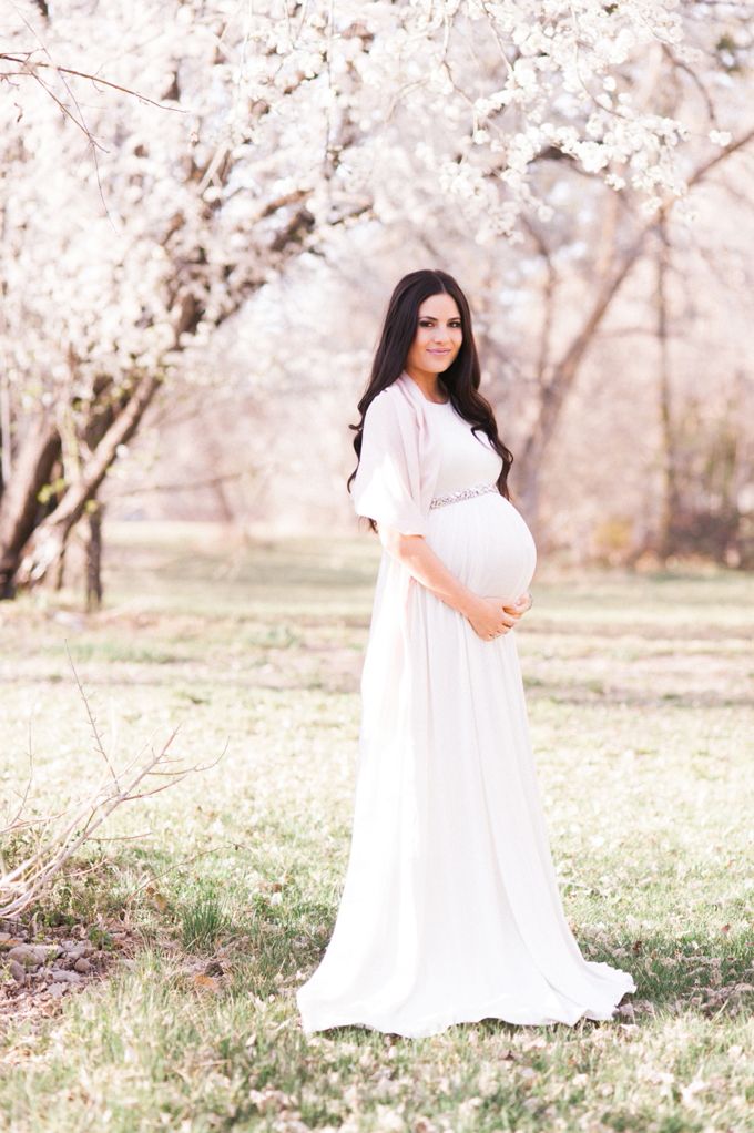 a pregnant woman in a white dress poses for a photo under the blossomy trees