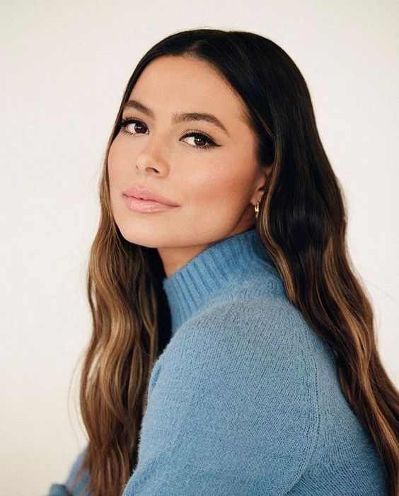 a woman with long brown hair wearing a blue turtle neck sweater and gold hoop earrings