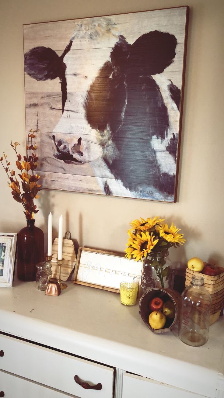 a painting hangs on the wall above a dresser with flowers and fruit in vases