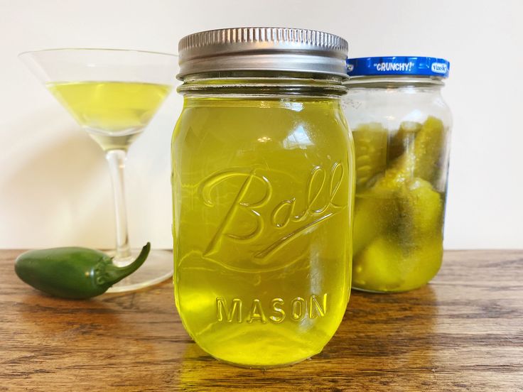 two mason jars filled with pickles next to a glass of wine and a green pepper