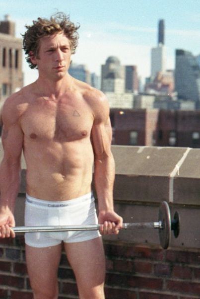 a shirtless man is holding a barbell on top of a building in the city