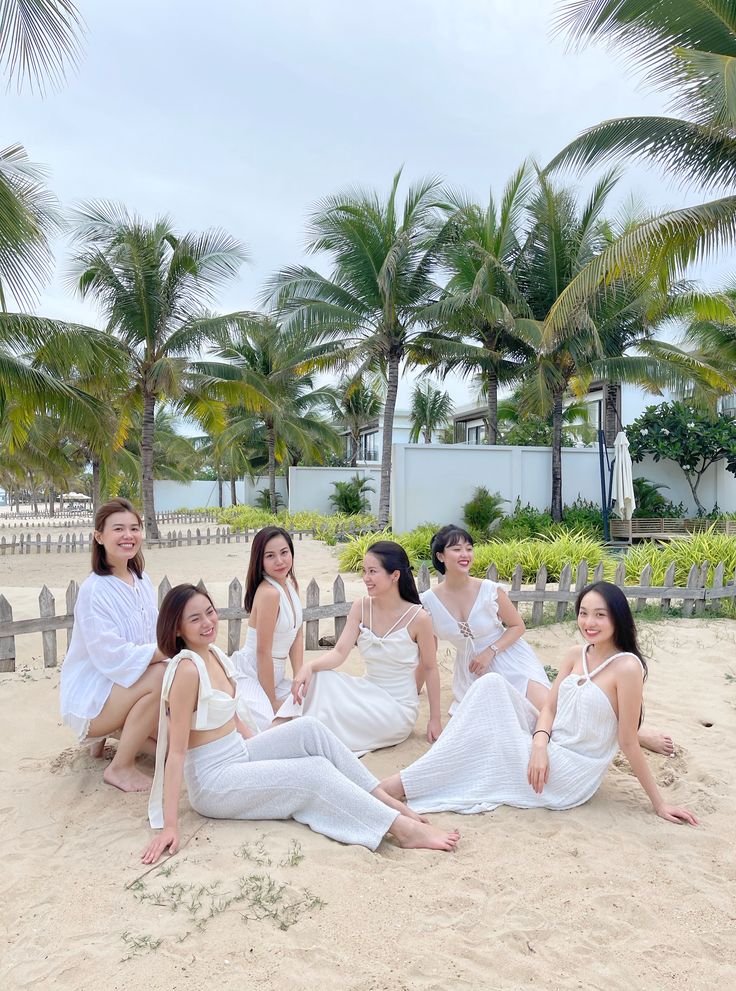 four women are sitting on the sand in white outfits and posing for a photo with palm trees behind them