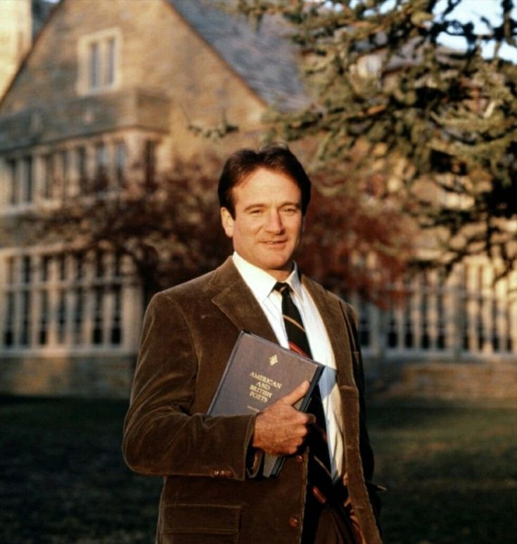 a man wearing a suit and tie holding a book in front of a large house