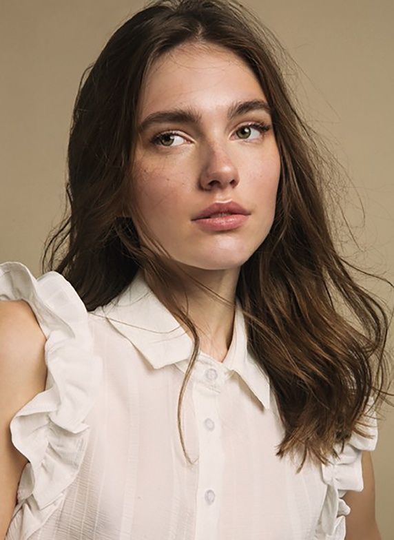 a woman with long brown hair wearing a white shirt and looking at the camera while standing in front of a beige background