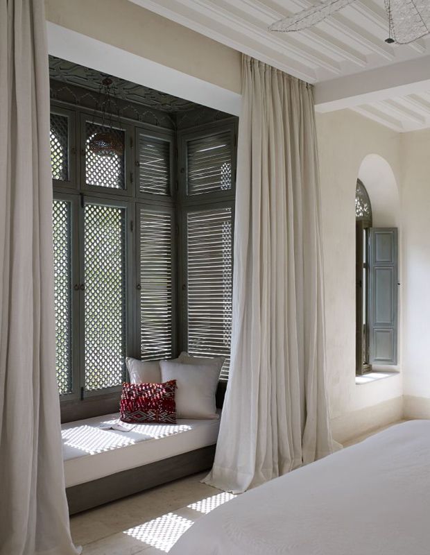 a white bed sitting next to a window covered in curtained drapes and pillows