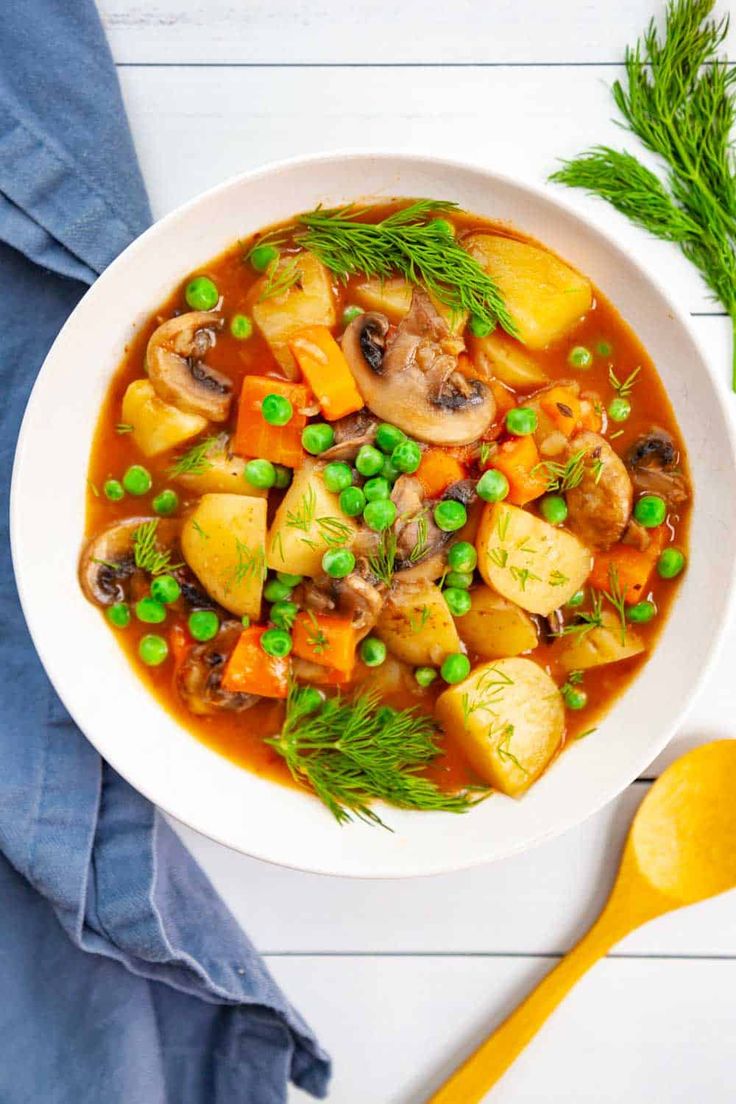 a white bowl filled with vegetable stew next to a yellow spoon and blue napkin on the side