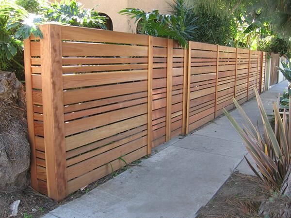 a wooden fence with plants growing on top of it next to a sidewalk and trees