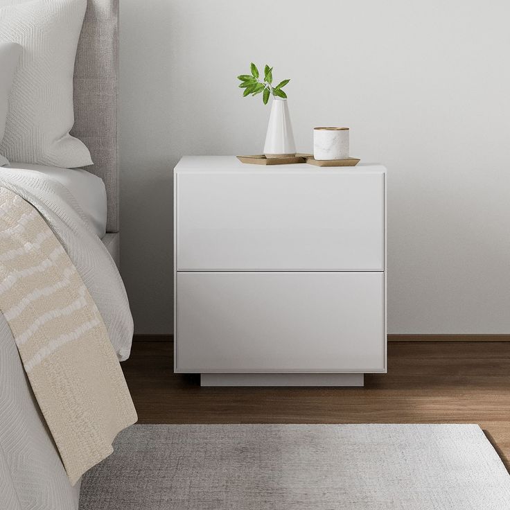 a white nightstand next to a bed in a room with hardwood floors and walls, along with a plant on top of the night stand