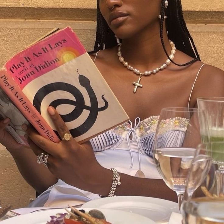 a woman sitting at a table with a book in her hands and wine glasses on the side