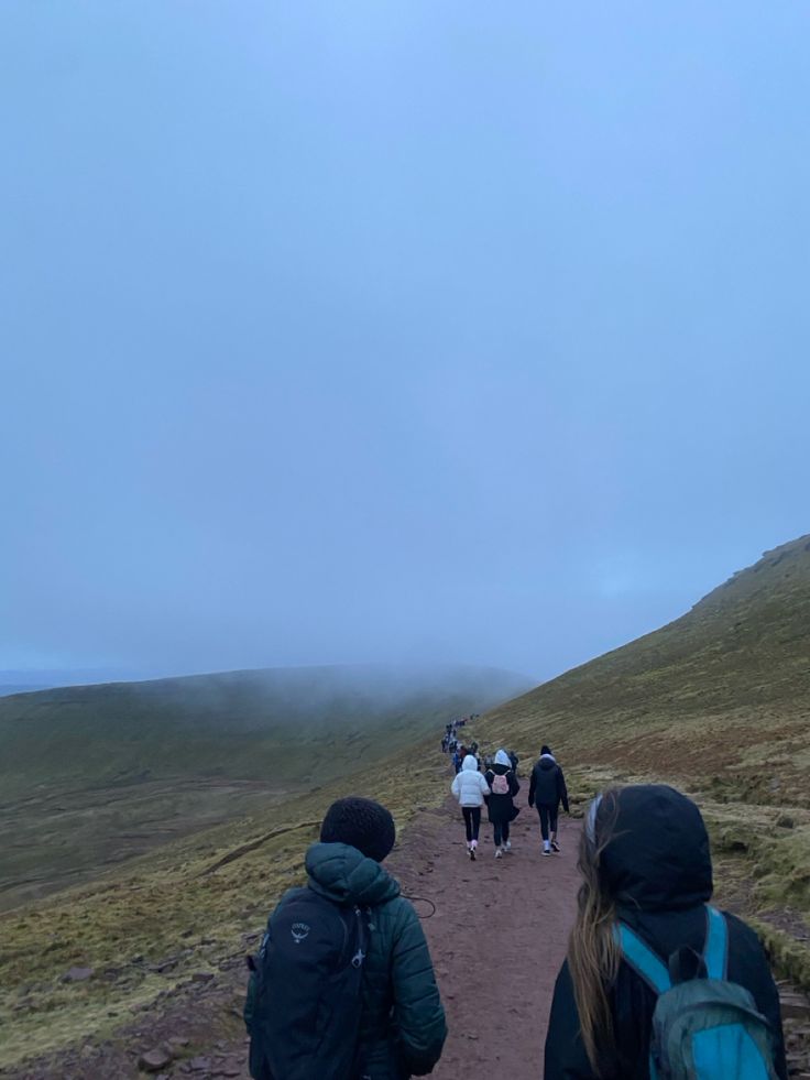 group of people walking up a dirt path in the mountains on a foggy day