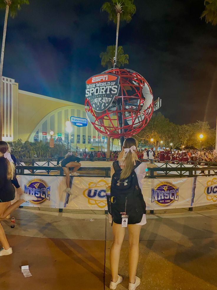 two women are standing in front of a sports sign at night with their hands behind their heads