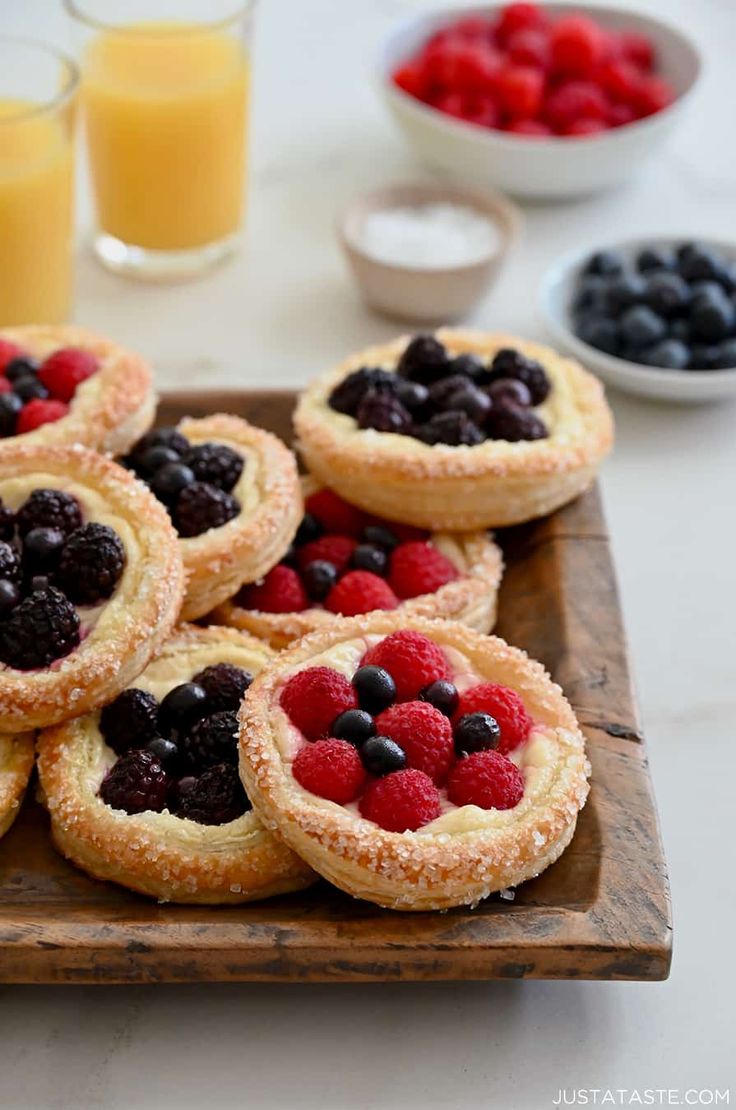 small pastries with berries and blueberries are on a wooden tray next to glasses of orange juice