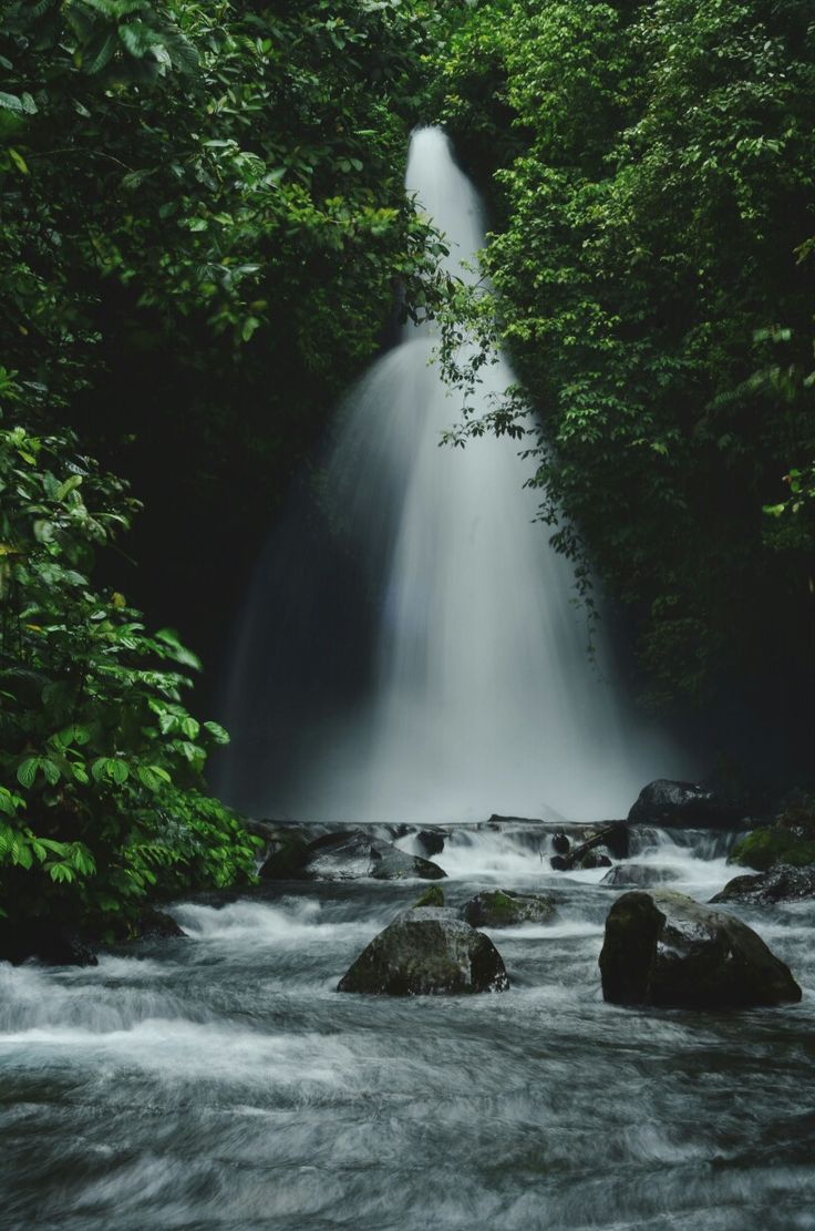 a large waterfall in the middle of a forest filled with rocks and greenery on either side