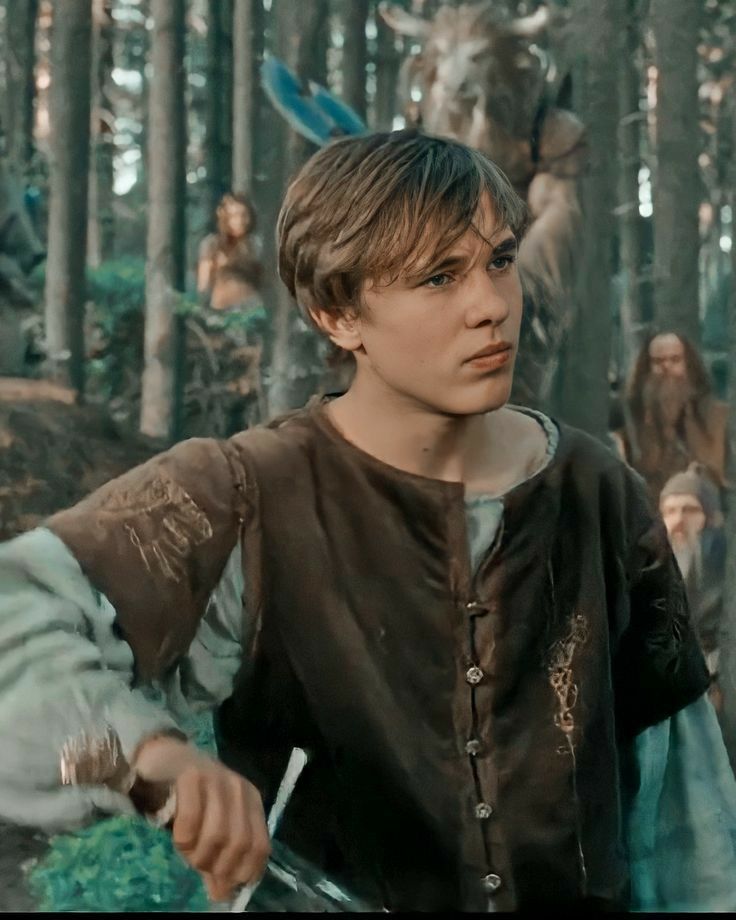 a young boy dressed in medieval clothing standing in the woods with other people behind him