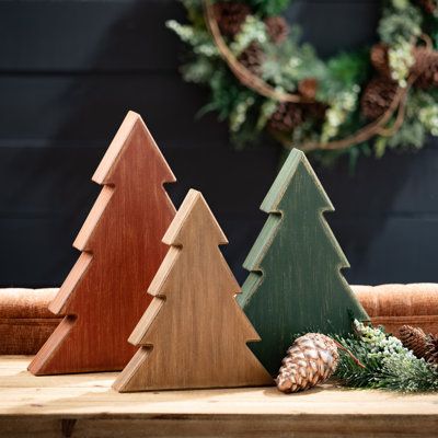 three wooden christmas trees sitting on top of a table next to a wreath and pine cones