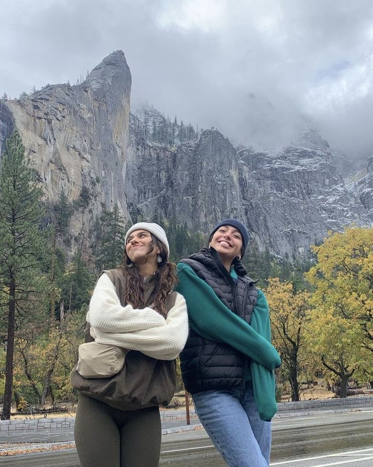 two women standing next to each other on the side of a road with mountains in the background