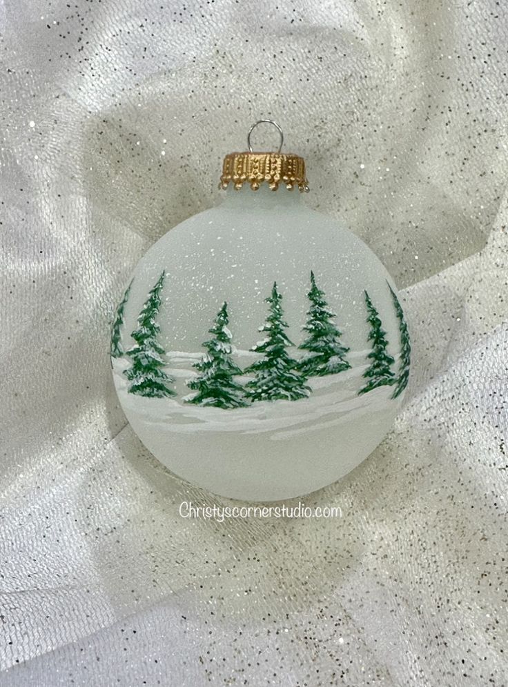 a glass ornament with trees painted on the front and bottom, sitting on a white fabric