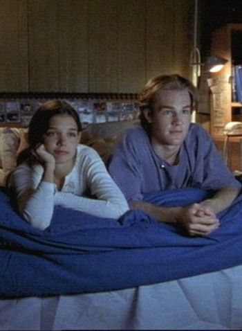 a man and woman laying on a bed with blue sheets in a room full of furniture